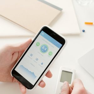 Close-up of hands using a smartphone app to monitor glucose levels with a digital glucometer on a table.
